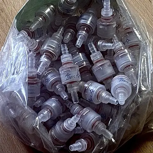 An open clear plastic bag filled with many clear barbed check valves on a wood table. The centermost check valve displays the company name "Air Logic"